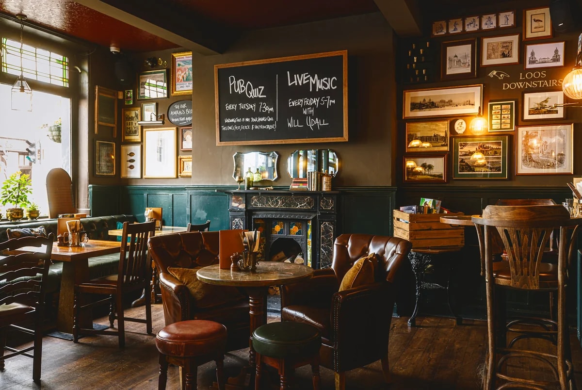 The image shows a cozy pub or bar with dark green walls, wooden floors, and a fireplace. The bar has a wooden table with a vase of flowers and a wooden bench. The walls display framed pictures and a chalkboard menu. The bar is well-lit, with a window allowing natural light. The scene is inviting and cozy, with a focus on the bar's ambiance and atmosphere.