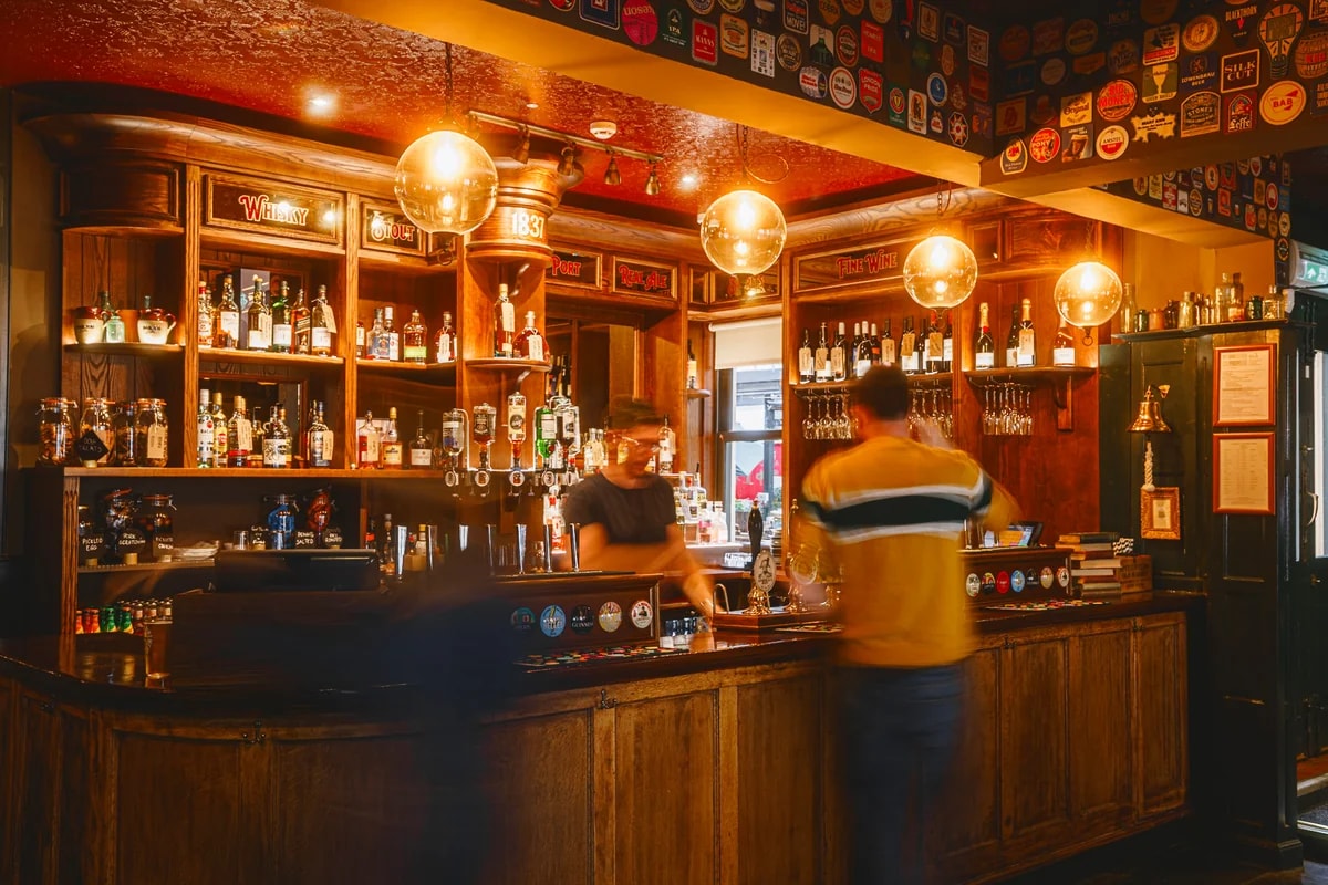 In a cozy bar, a wooden bar with a red and white patterned ceiling and gold chandeliers casts a warm glow. Three shelves display various liquor bottles and glasses, while a bar staff in a yellow and black striped shirt stands behind the counter. The scene is bathed in soft, warm light, creating a welcoming atmosphere.