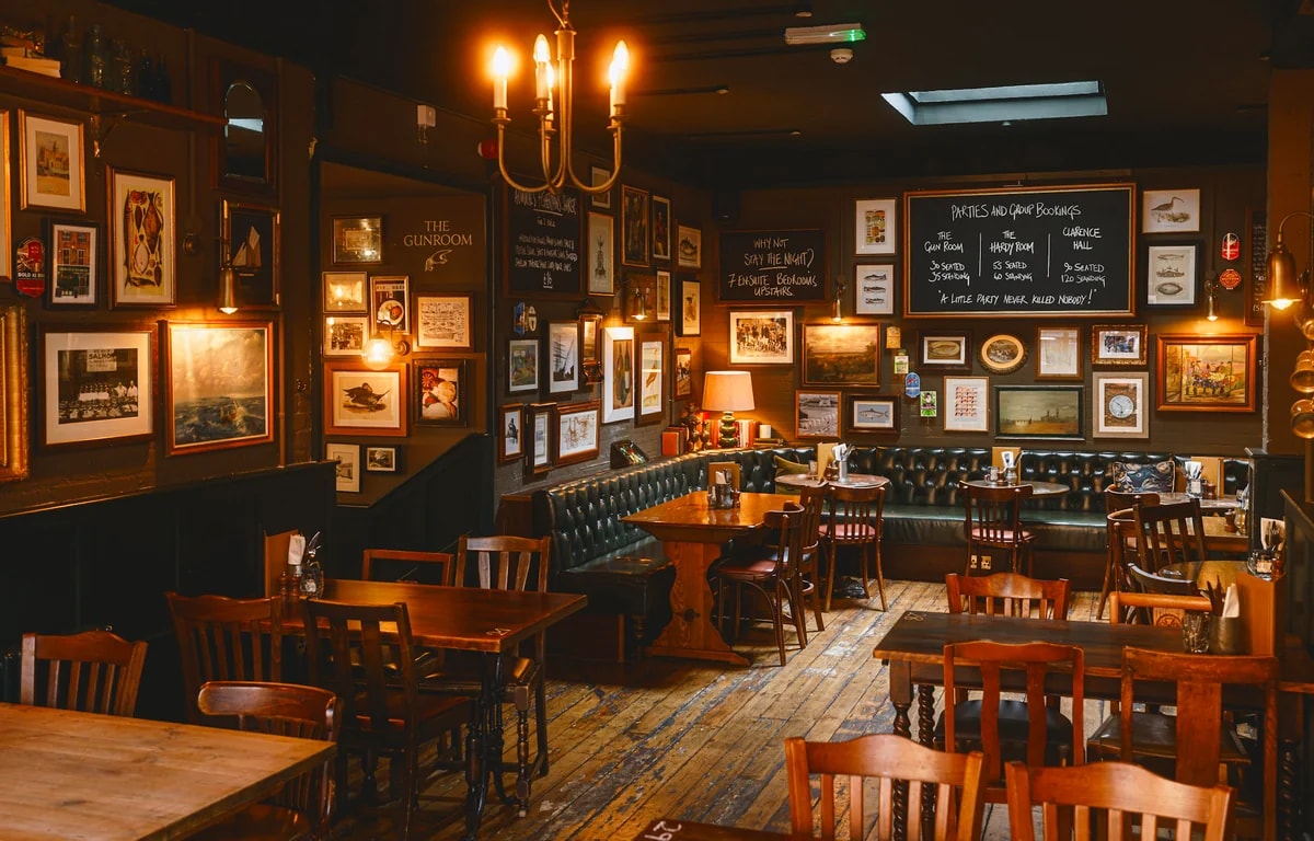 The image shows a cozy, dimly lit bar with dark wood floors and a wooden bar counter. The walls are adorned with framed pictures and posters, and a chalkboard menu is displayed. The bar is filled with wooden tables and chairs, and a green leather couch is in the background. The atmosphere is warm and inviting, with a chandelier casting a soft glow.