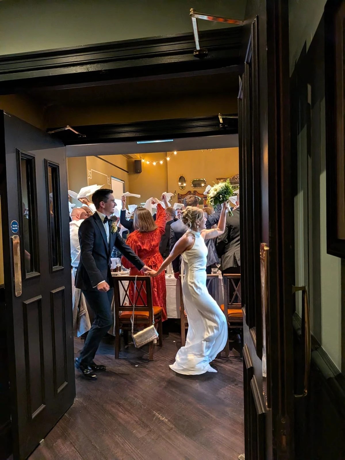 A joyous wedding scene unfolds in a dimly lit room, with a bride in a white dress and a groom in a black suit. They exit a wooden door, surrounded by guests in vibrant attire. The room is decorated with string lights and a red banner, creating a festive atmosphere. The image captures the joyous moment of the couple's journey into their new life.