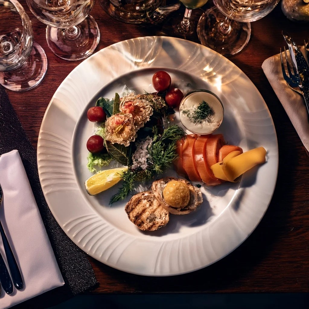 A white plate with a variety of food items is at the center of a dark wooden table, surrounded by a black napkin, fork, knife, and wine glasses. The plate features salmon, cheese, cherry tomatoes, and a small white sauce. The arrangement creates a visually appealing and balanced scene.