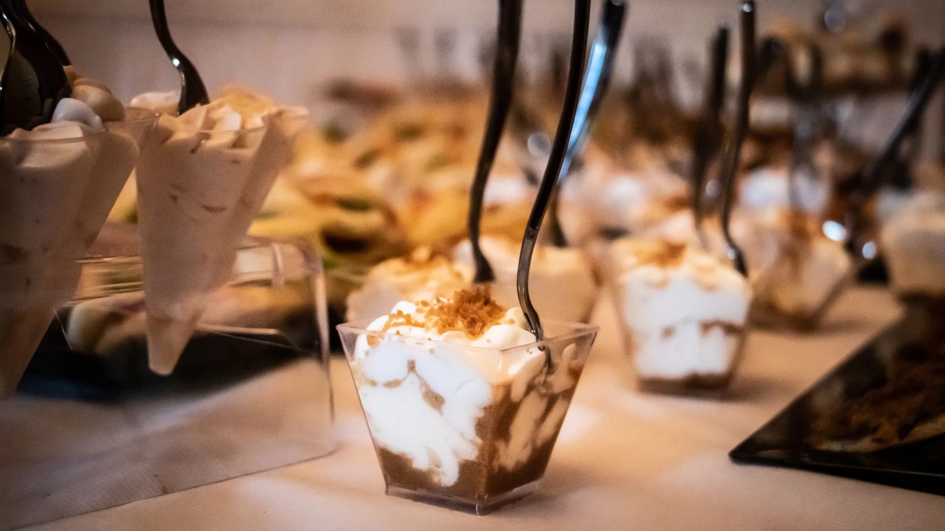 The image features a table with a white tablecloth, displaying a variety of desserts in small glass cups. The desserts include vanilla ice cream, chocolate mousse, and a dessert with whipped cream and caramel sauce. The background is blurred, emphasizing the desserts and their presentation. The image captures a moment of anticipation, with the desserts ready to be enjoyed.
