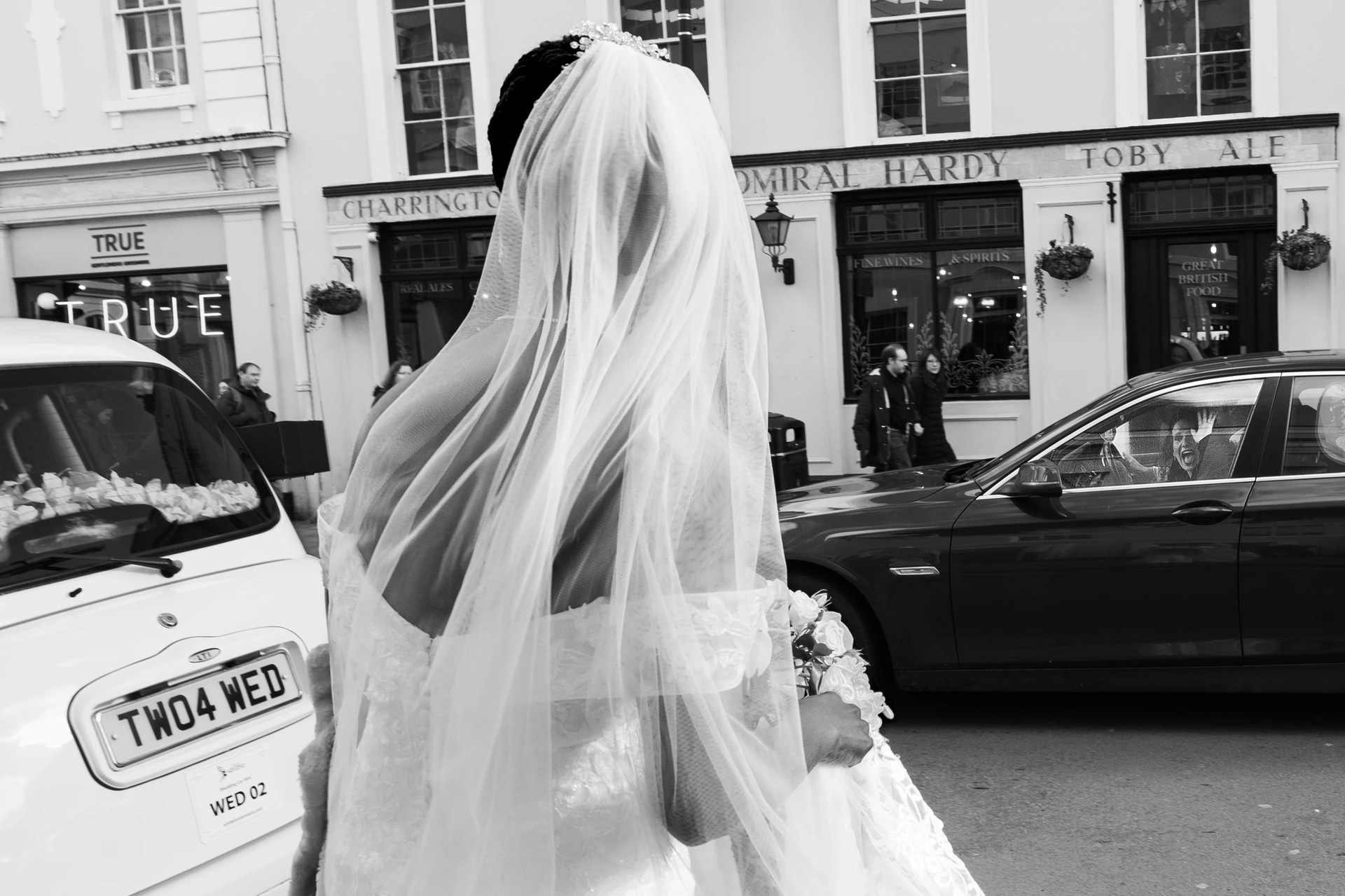 A bride in a white wedding dress and veil, holding a bouquet of flowers, walks away from the camera on a bustling city street. The photo is taken from a low angle, emphasizing the bride's grandeur. The background features a "The Coffee Shop" and "The Bar" establishments, with a parked car and a "Kings" sign. The image captures a moment of joy and celebration in a city setting.