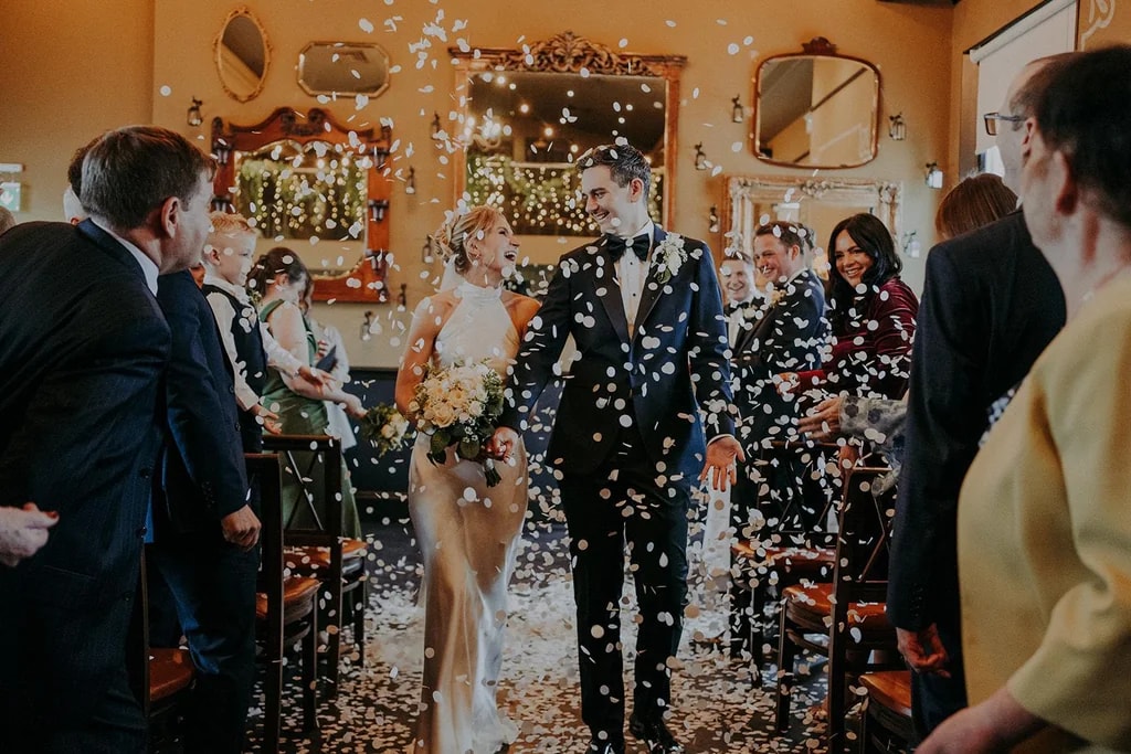 A newlywed couple, dressed in formal attire, exits a room with a shower of confetti. The bride holds a bouquet of flowers, and the groom wears a tuxedo. They walk towards the camera, surrounded by guests in the background. The room has a warm, festive atmosphere with a mirror and chandelier.