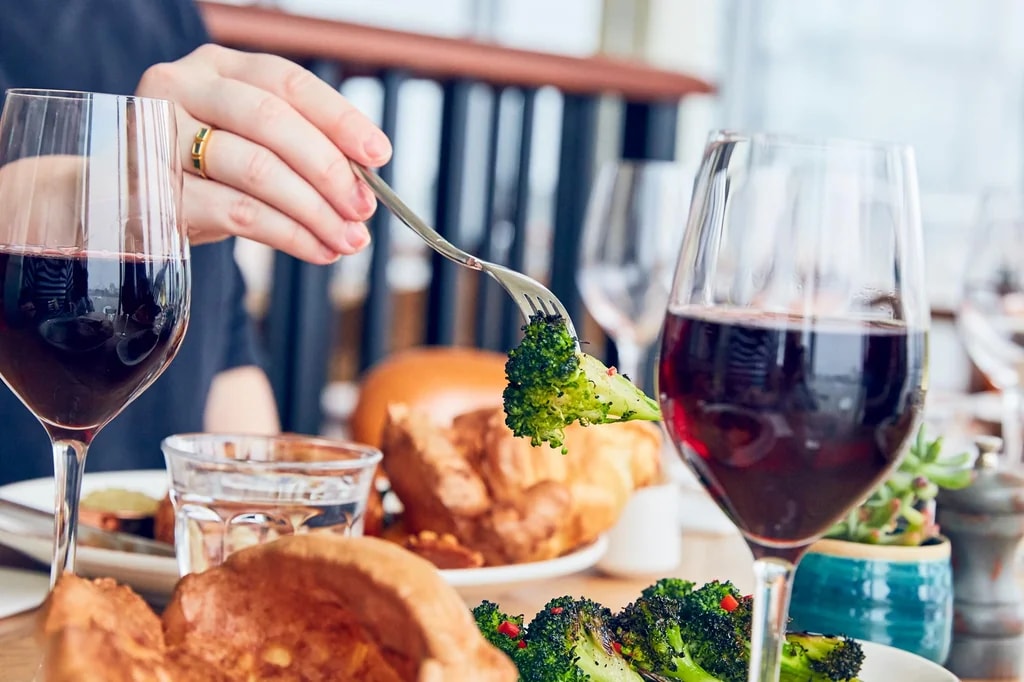A person is seated at a table, enjoying a meal with a large plate of food, including a piece of broccoli. Two glasses of red wine are nearby, and a small potted plant adds greenery. The blurred background suggests a restaurant or dining area. The scene is warm and inviting, with the main subject and key elements creating a sense of anticipation for the meal.