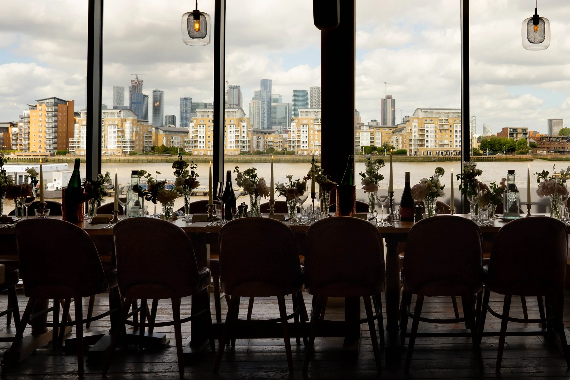 A modern restaurant with large windows offers a panoramic view of the city skyline. The restaurant features wooden tables and chairs, white tablecloths, and wine glasses. The city skyline is visible through the windows, with tall buildings and a river in the background. The restaurant is well-lit and has a modern aesthetic.
