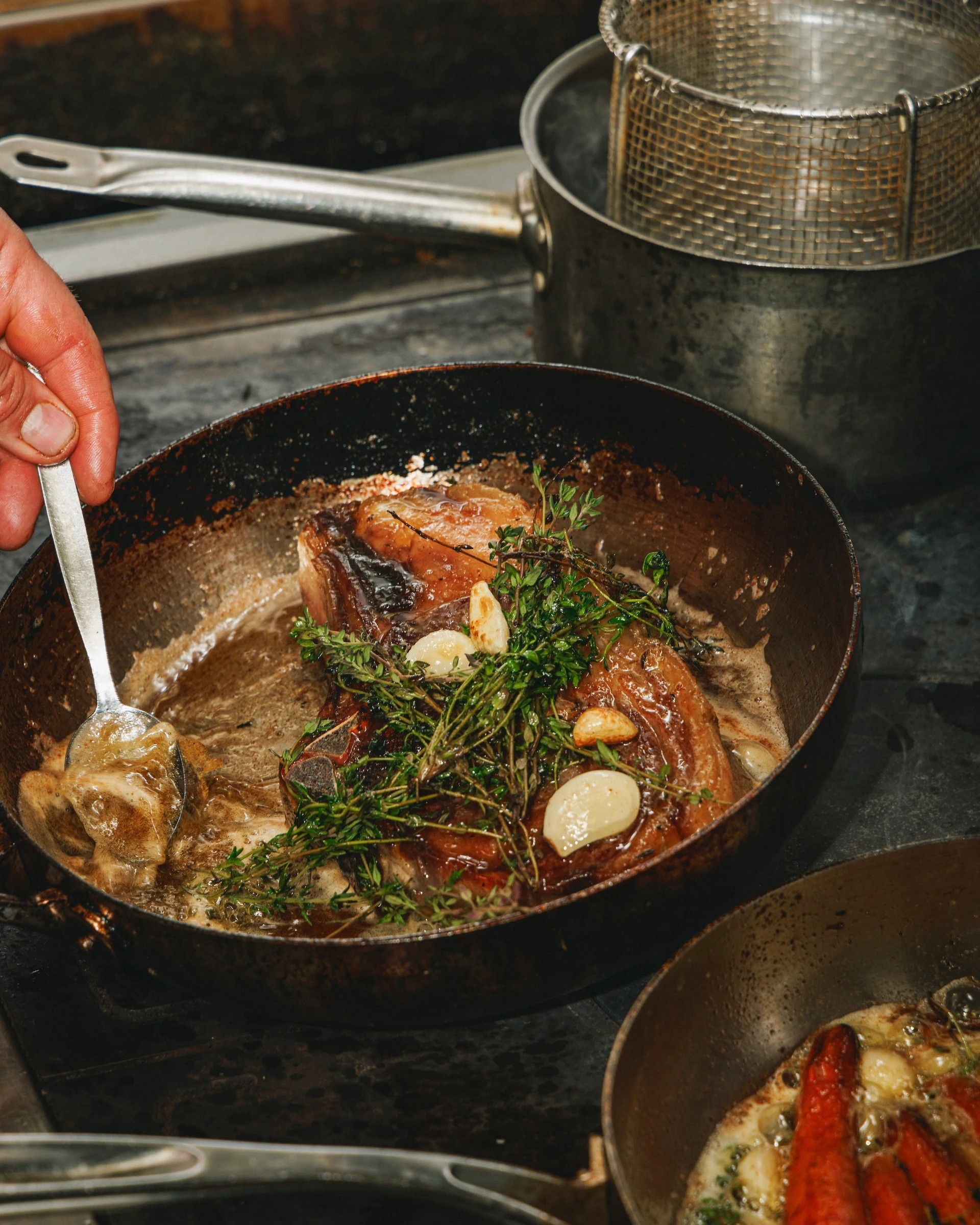 A hand is stirring a pan filled with a hearty meal, including meat, vegetables, and herbs, on a black stove. A silver pot and a metal container are nearby, and a silver pot is on the stove. The scene is warm and inviting, with a focus on the cooking process.