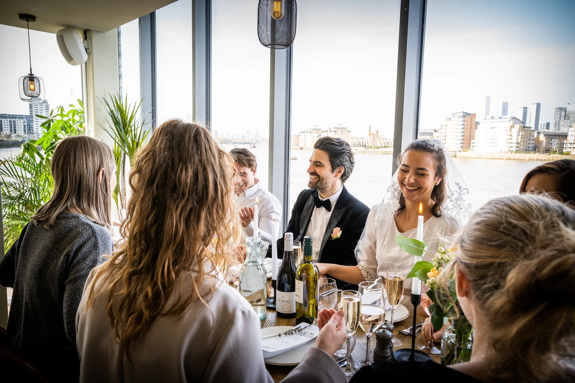 A lively restaurant scene features a group of people enjoying a meal at a table with a white tablecloth, surrounded by a large window offering a view of the city skyline. The table is adorned with wine bottles, a candle, and a flower arrangement, while the people are engaged in conversation and laughing. The atmosphere is warm and inviting, with a man in a black tuxedo and a woman in a white sweater. The cityscape in the background adds depth to the scene.