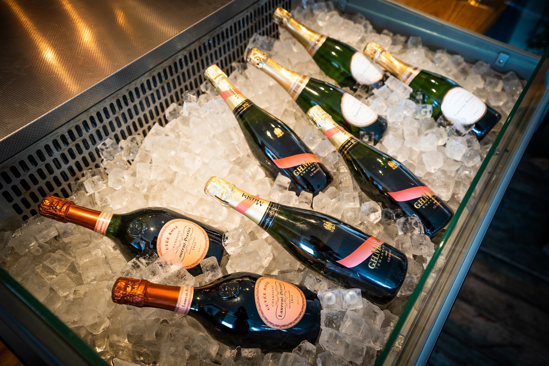Six champagne bottles, each with a gold label and a pink label, are arranged in a diagonal line on a metal tray with ice cubes. The bottles are green with gold labels and pink labels, and the tray is on a wooden surface. The scene is set against a warm, ambient light, creating a festive atmosphere.
