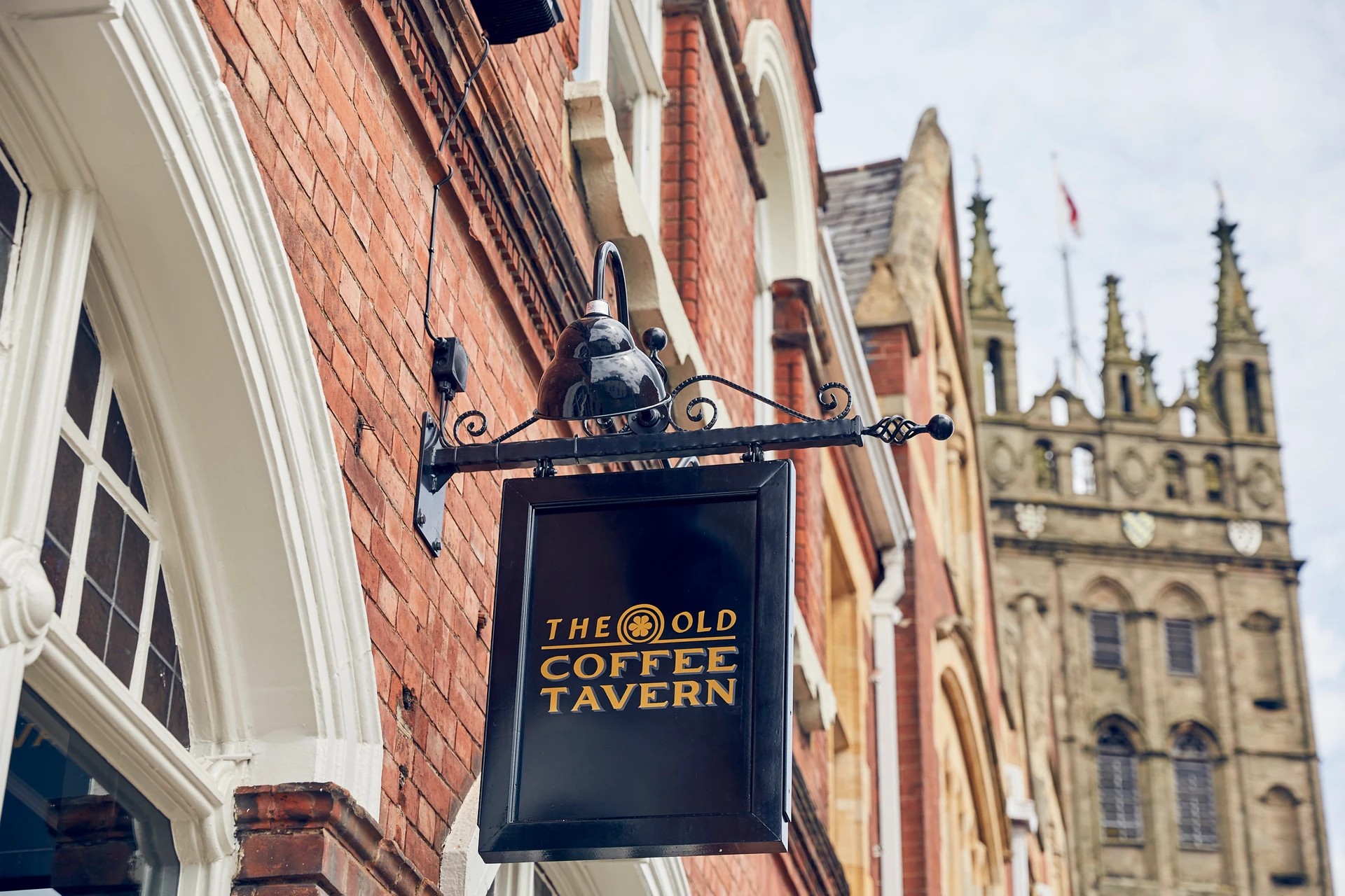 The image features the "The Old Bank" sign on a black metal lantern, hanging from a brick building with a white archway. The building has a red brick facade and a clock tower in the background. The sky is overcast, and the scene is serene and inviting.