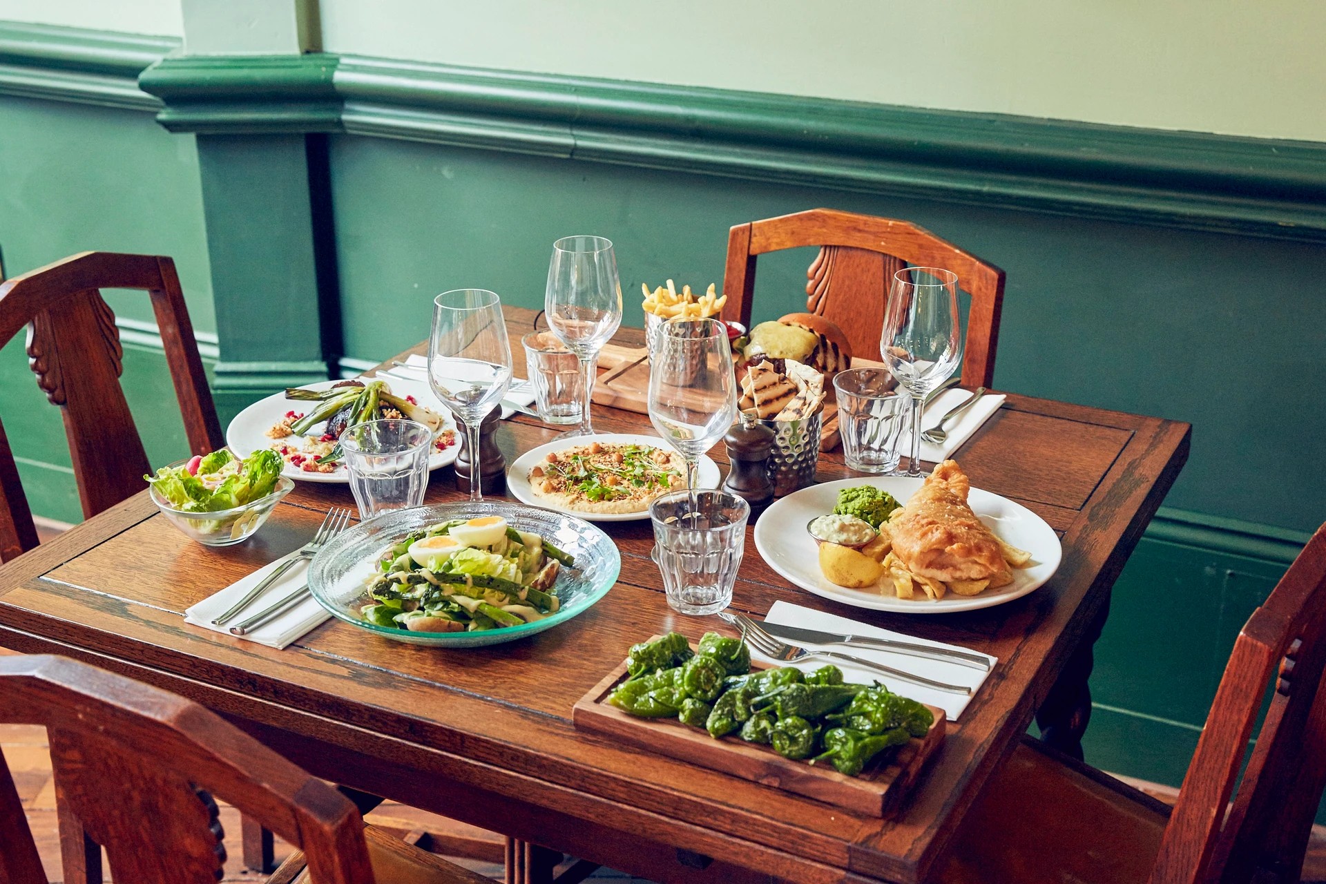 A wooden dining table is set for a meal, with a variety of dishes and utensils arranged. The table features a basket of bread, a basket of salad, a plate of fried chicken, a plate of vegetables, and a plate of French fries. The table is surrounded by wooden chairs, and the background is a green wall with a window. The scene is inviting and ready for a meal.
