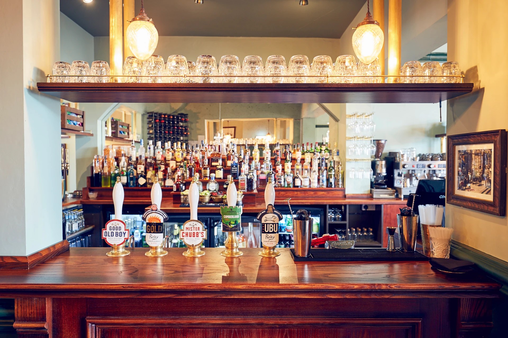 The image features a well-organized bar with a wooden counter, white wall, and gold-framed mirror. The counter is filled with various liquor bottles, including whiskey, gin, and vodka. A black coffee machine and a wooden bar stool are also present. The background reveals a wall with a framed picture and a window, creating a cozy atmosphere. The arrangement of objects and lighting contribute to the overall ambiance of the bar.