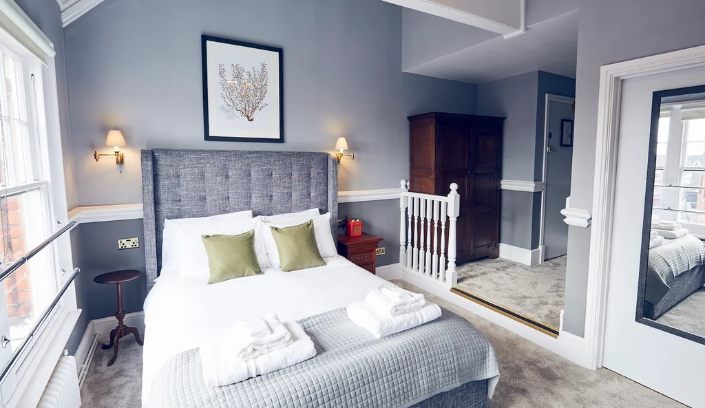 The image shows a serene bedroom with a large gray bed, white linens, and green and white pillows. Two lamps with warm light create a cozy atmosphere. A wooden nightstand with a red vase and a mirror above the bed add sophistication. A wooden wardrobe and a white rail on the floor complete the room's design. The walls are painted in soothing shades of blue, and a framed art piece adds a touch of elegance. The room is well-organized and inviting, with a harmonious color palette and thoughtful design.