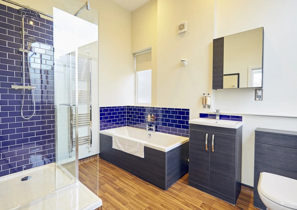 The image shows a modern, well-lit bathroom with a white bathtub, silver faucet, and blue tiles. A white toilet is to the right, and a glass shower enclosure is on the left. A wooden cabinet with a silver faucet is in front of the bathtub, and a mirror above the bathtub reflects the room's color scheme. The floor is wooden, and the walls are painted in a soothing yellow. The bathroom is clean, organized, and functional.
