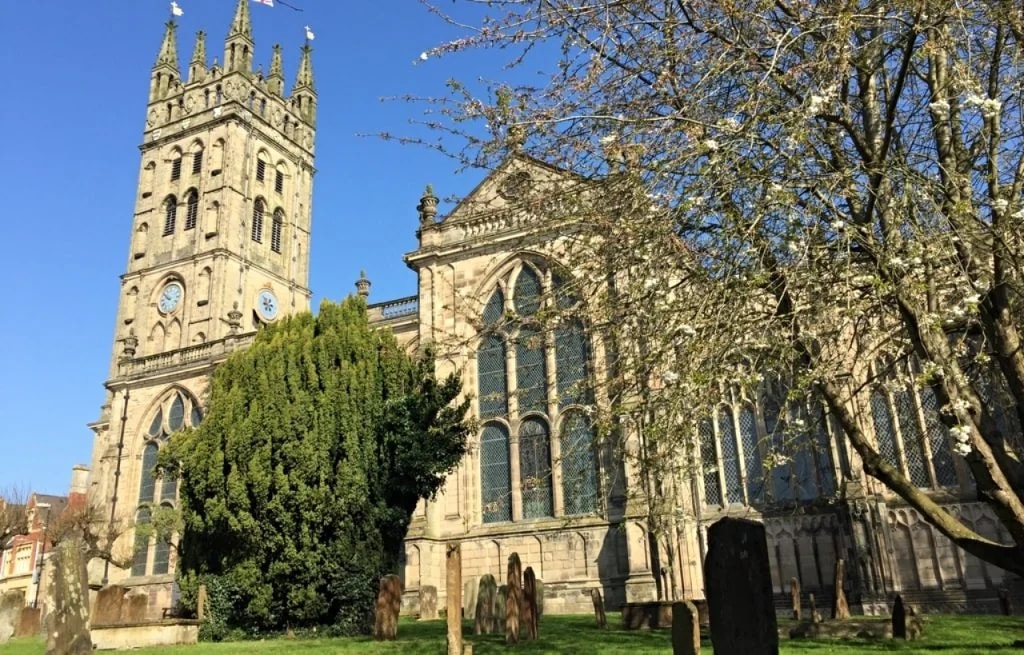 The image features a Gothic church with a tall, gray stone tower and two smaller towers, all adorned with arched windows and pointed roofs. The church is surrounded by a cemetery with gravestones and a tree with white flowers. The sky is clear blue, and the church is centrally located in the frame. The image captures a serene, tranquil scene with architectural details and natural elements.