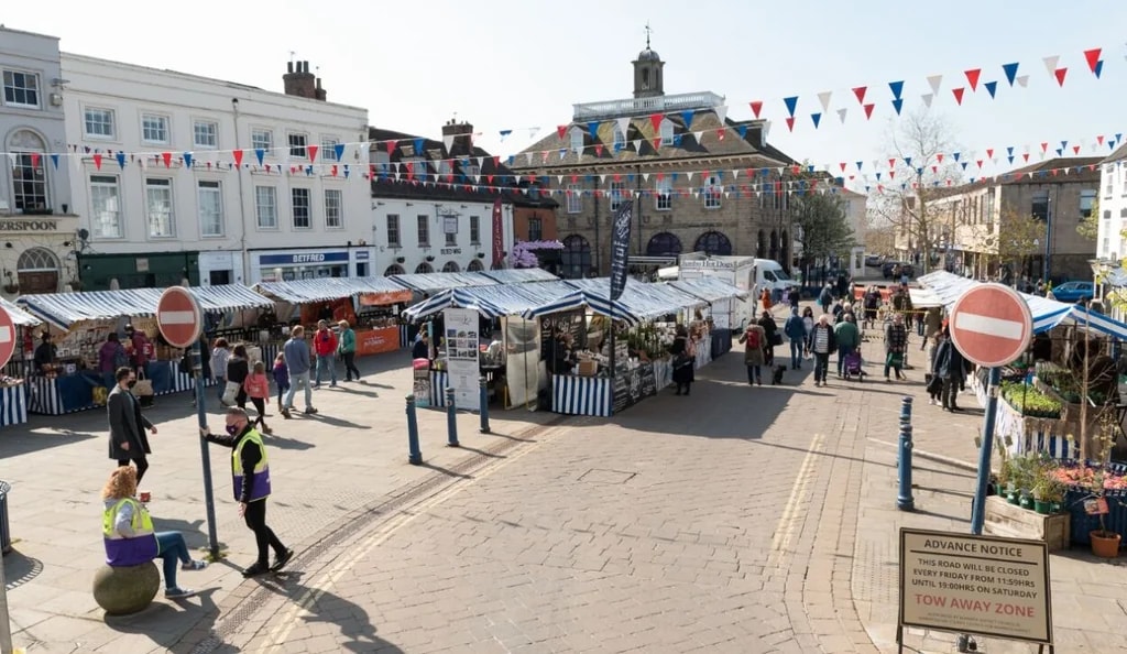 The image shows a bustling market in a town square, with white buildings and blue and red flags. The square is filled with colorful stalls, selling various items. People are browsing and shopping, while a clock tower stands in the background. The scene is lively and vibrant, showcasing community engagement and local culture.