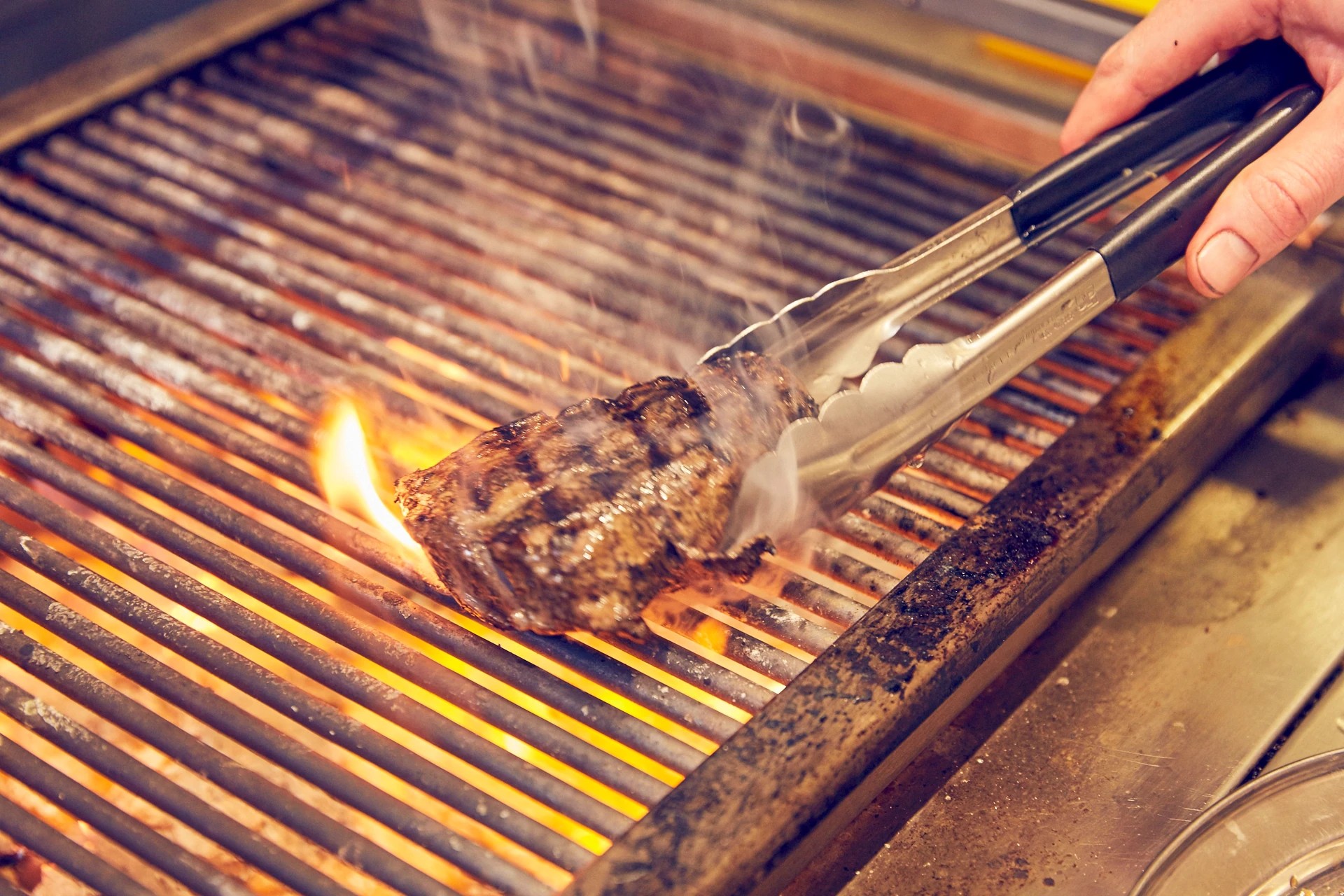A hand skillfully handles a steak on a grill, with a tongs held over the steak and the grill's flames dancing around it. The background is blurred, emphasizing the steak and grill. The image captures a moment of culinary artistry and precision.
