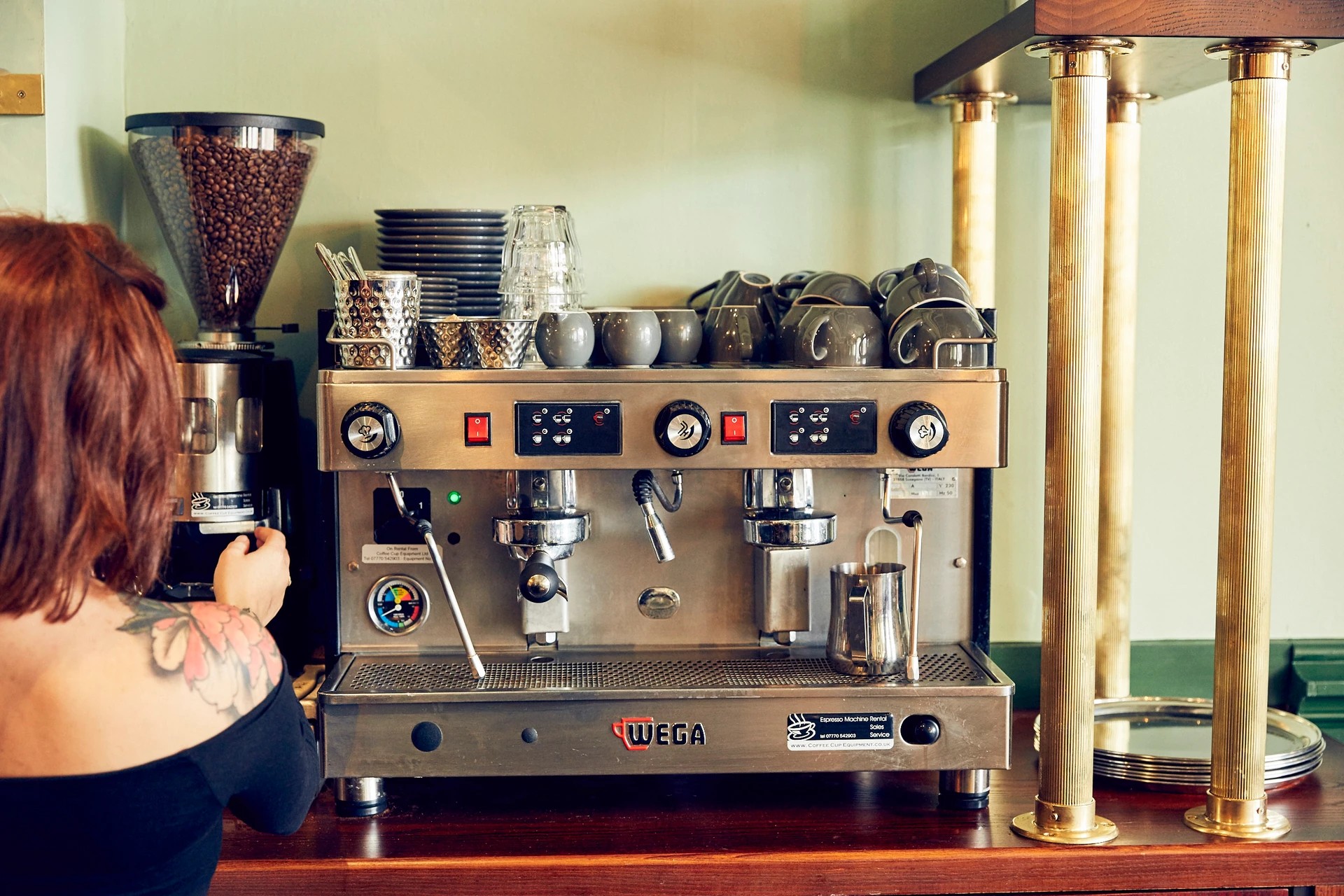 A woman with red hair and tattoos is in a coffee shop, holding a glass of water and looking at a large, silver Oster coffee maker with red and black buttons. The machine is on a wooden counter, with a stack of coffee cups and a coffee grinder nearby. The background features a green wall and a gold pillar, creating a warm and inviting atmosphere.