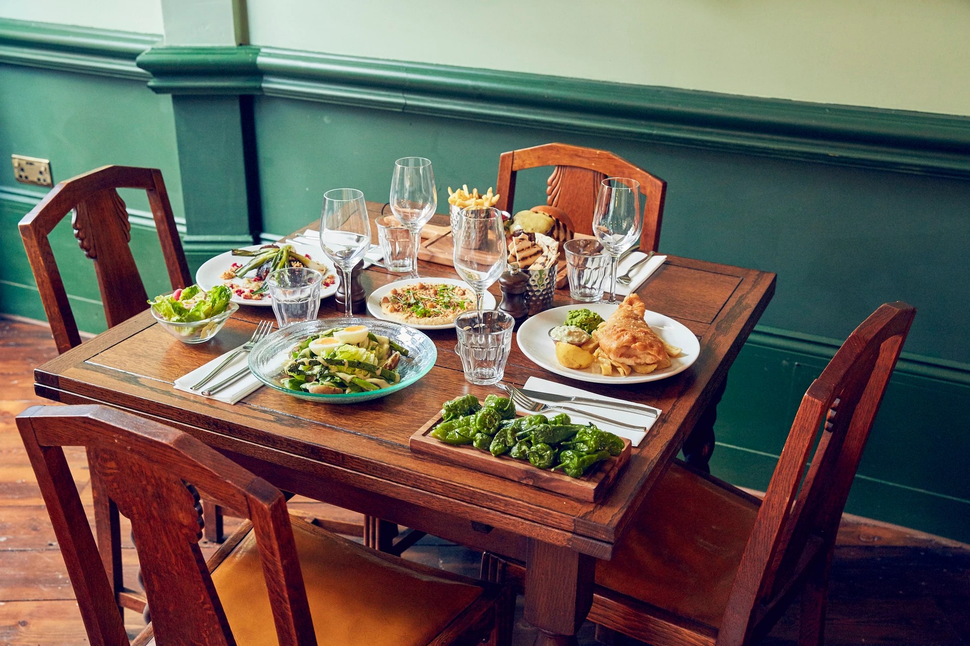 The image depicts a beautifully set dining table in a restaurant, with a wooden tablecloth and four wooden chairs. The table is adorned with six plates of food, including a large salad, a fried chicken, a sandwich, and a baked potato. Six glasses of water and six wine glasses are also present. The table is surrounded by green walls and features a white electrical outlet. The scene is inviting and suggests a meal is about to be enjoyed.