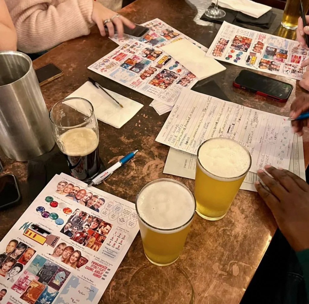 In a lively bar, a wooden table is filled with various items, including two half-full glasses of beer, a silver pitcher, a black cell phone, and a white napkin. The scene is filled with people engaged in conversation, creating a dynamic atmosphere.