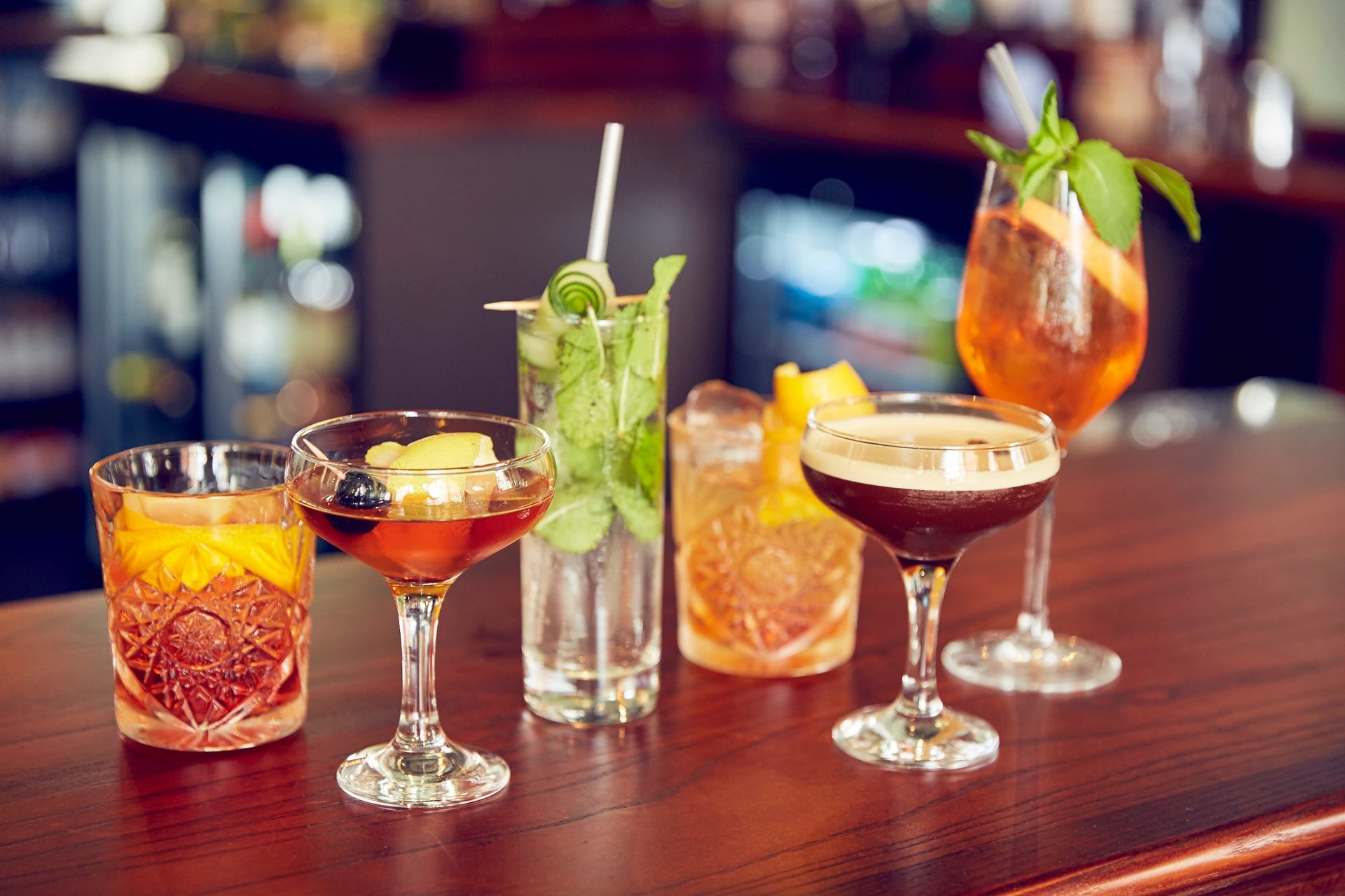 The image features a wooden bar with six glasses filled with various drinks, each with a straw and garnish. The glasses are arranged in a row, with the largest glass on the left and the smallest on the right. The background is blurred, focusing on the bar and drinks. The image is a snapshot of a moment in a bar, with the objects' positions and actions suggesting a lively atmosphere.
