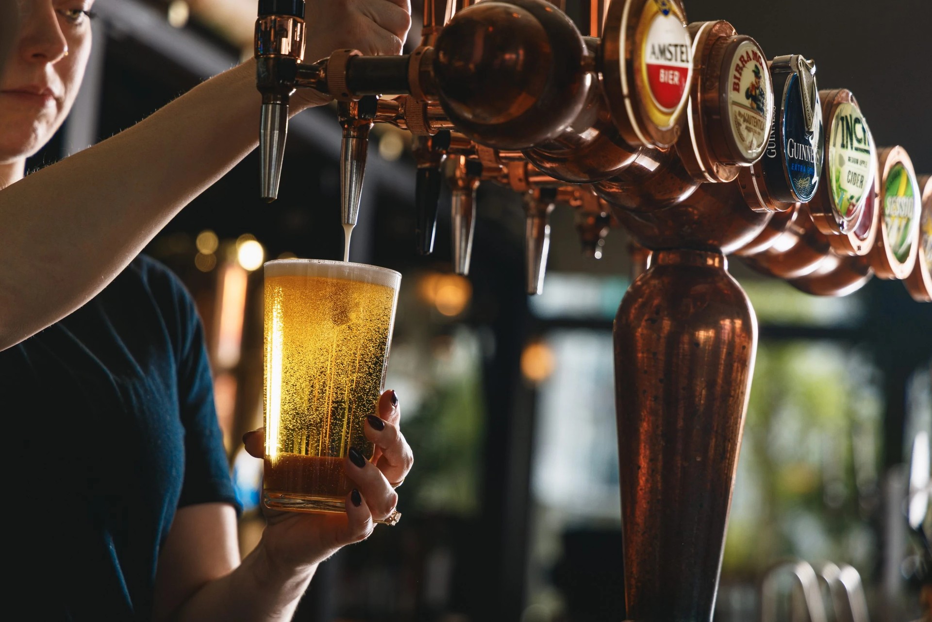 A person in a blue shirt is pouring beer from a tall glass into a wooden beer tap, with six beer bottles arranged in a row behind them. The background is blurred, focusing on the beer tap and person.