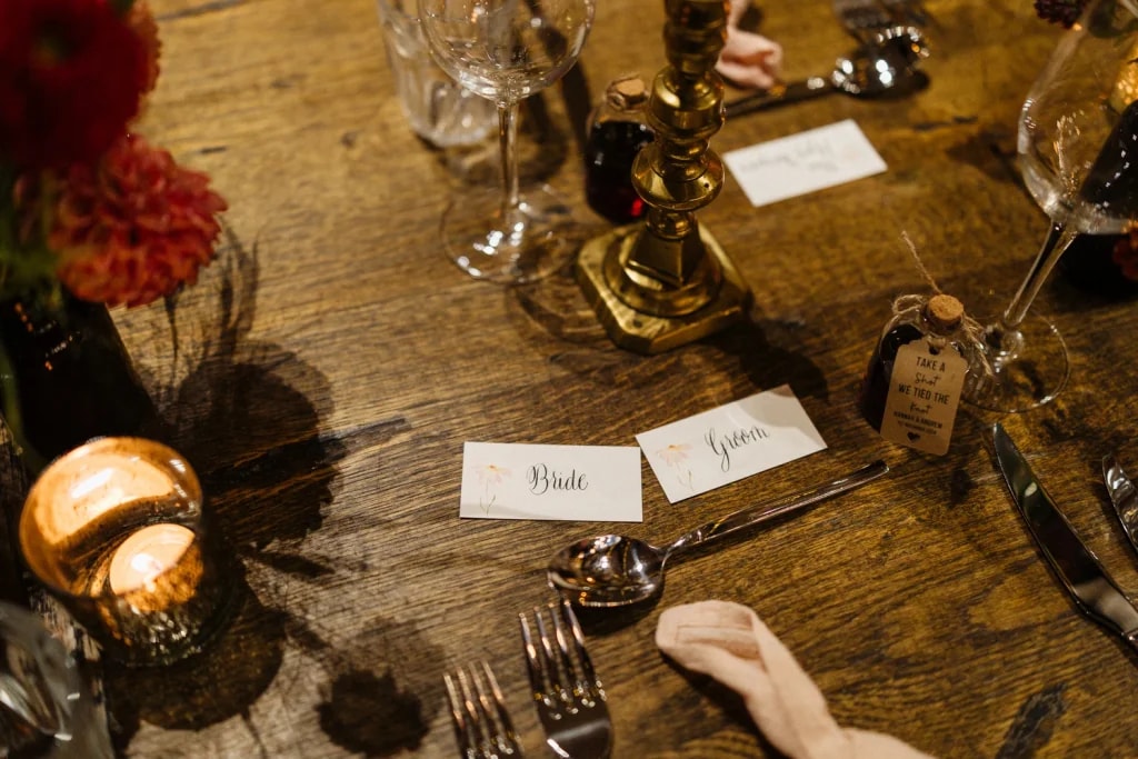 The image shows a beautifully set table for a formal dining event, with a wooden table, silverware, and candle. The table is adorned with a gold candle holder, a candle, and a vase of red and orange flowers. The arrangement suggests a well-thought-out table setting for a special occasion.