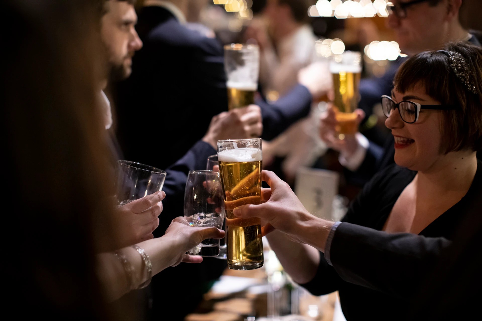 A lively bar scene features a group of people in the foreground, each holding a glass of beer and raising it in a toast-like gesture. The background is blurred, emphasizing the foreground activity. The image captures a moment of camaraderie and celebration.