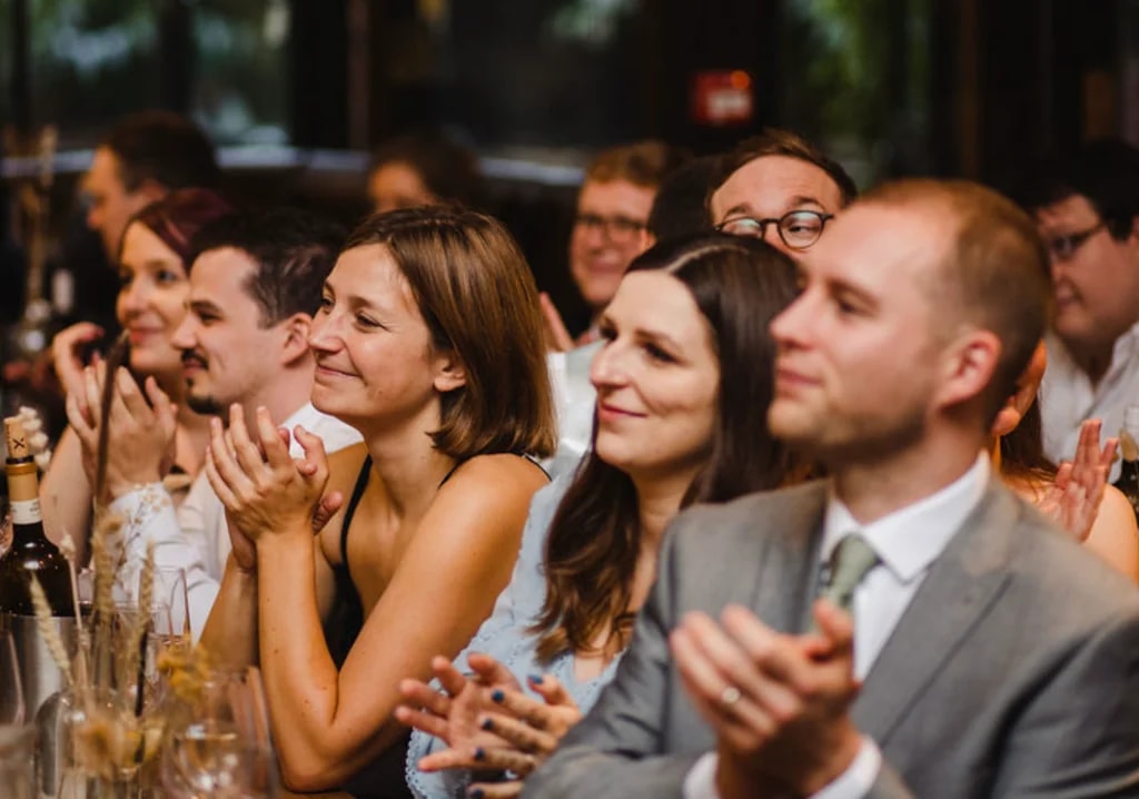 A lively wedding reception scene features a group of people, including a man in a gray suit and a woman in a black dress, both clapping and smiling. The background shows a bar with wine bottles and glasses, and a red fire alarm. The atmosphere is festive and joyful.