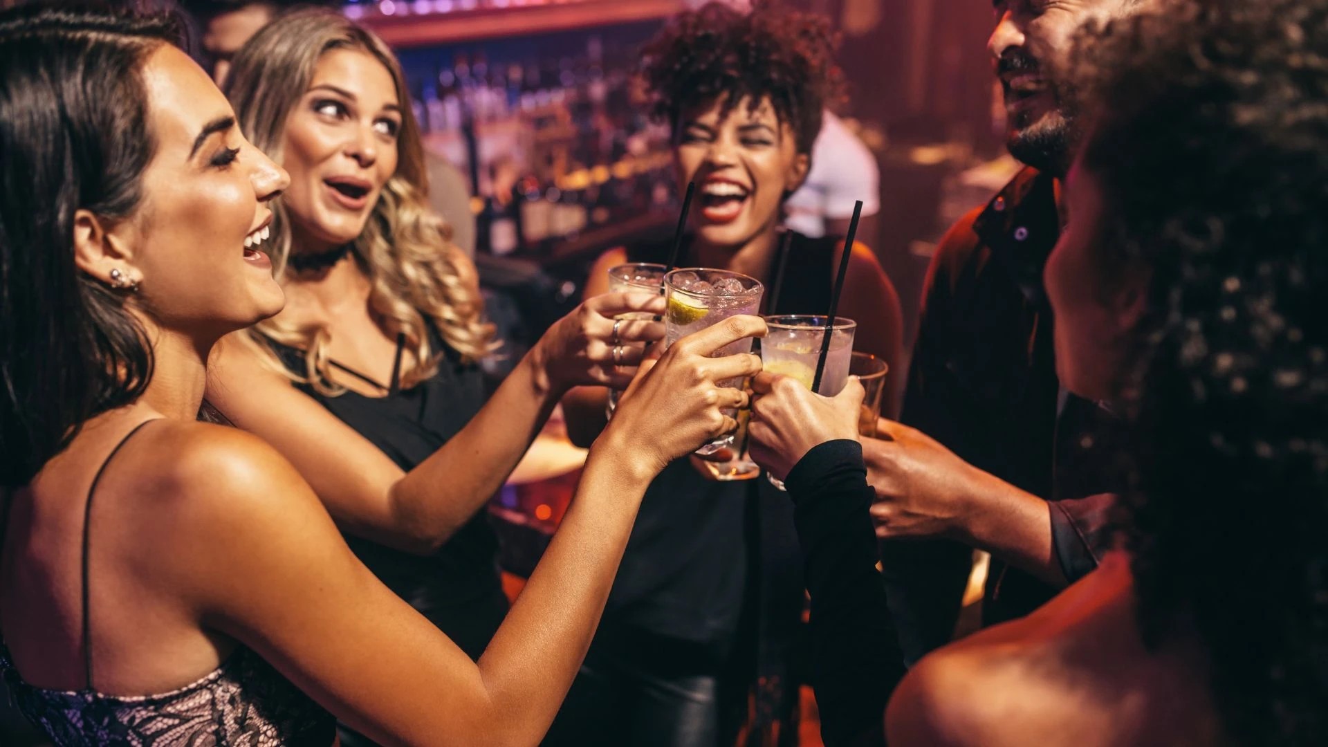 A lively bar scene features a group of women in vibrant attire, celebrating with a cocktail. The woman in the center holds a glass with a lime wedge and a straw, while others hold their own glasses. The background is a bustling bar with various drinks and a barista, creating a lively atmosphere.