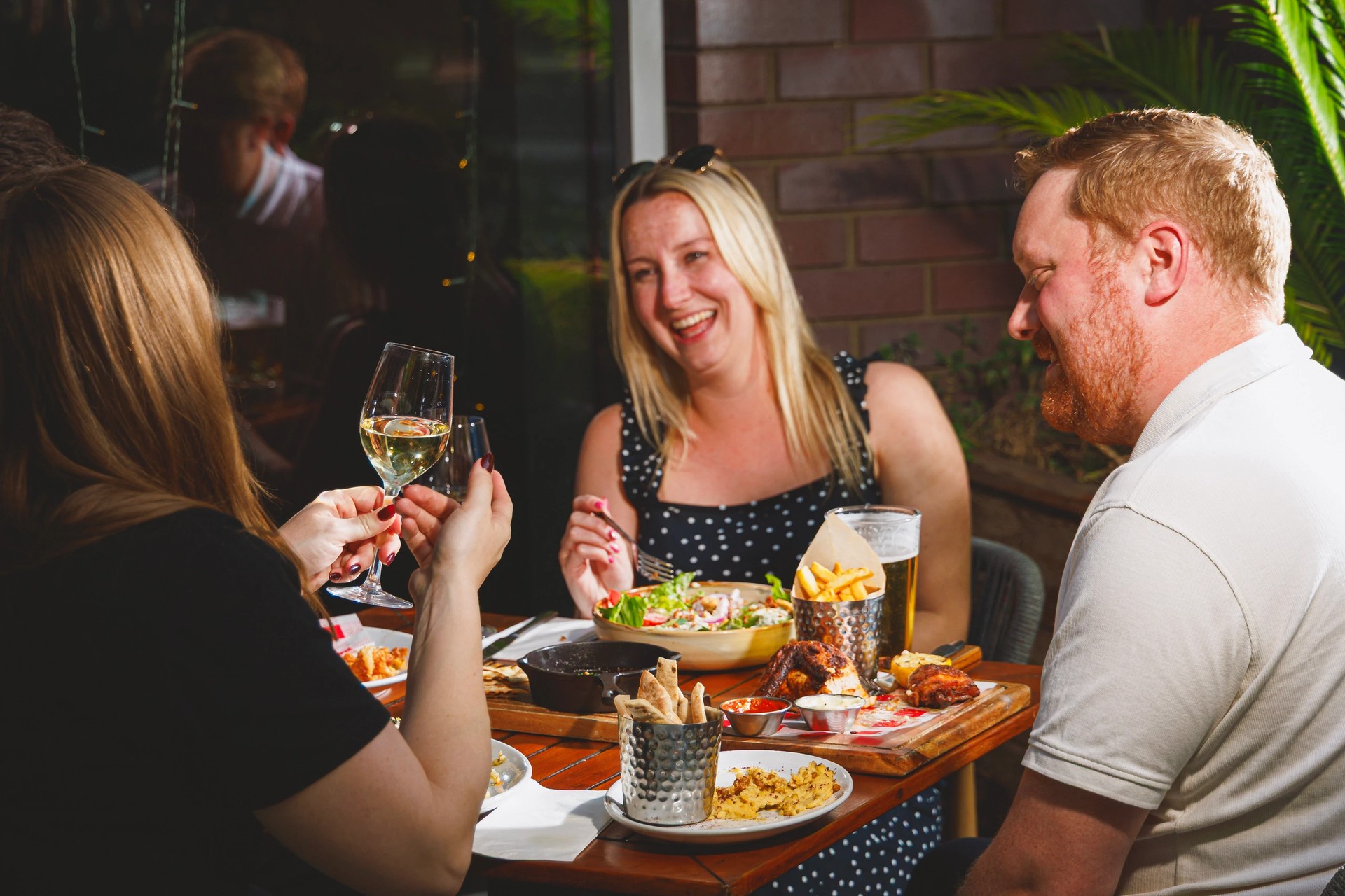 A lively gathering of friends and family at a rustic wooden table, surrounded by a brick wall and a window, enjoying a meal and conversation. The table is adorned with various dishes, glasses, and a white bowl of salad, while the group is engaged in animated conversation and laughter. The scene exudes warmth and camaraderie, capturing a moment of shared enjoyment and friendship.