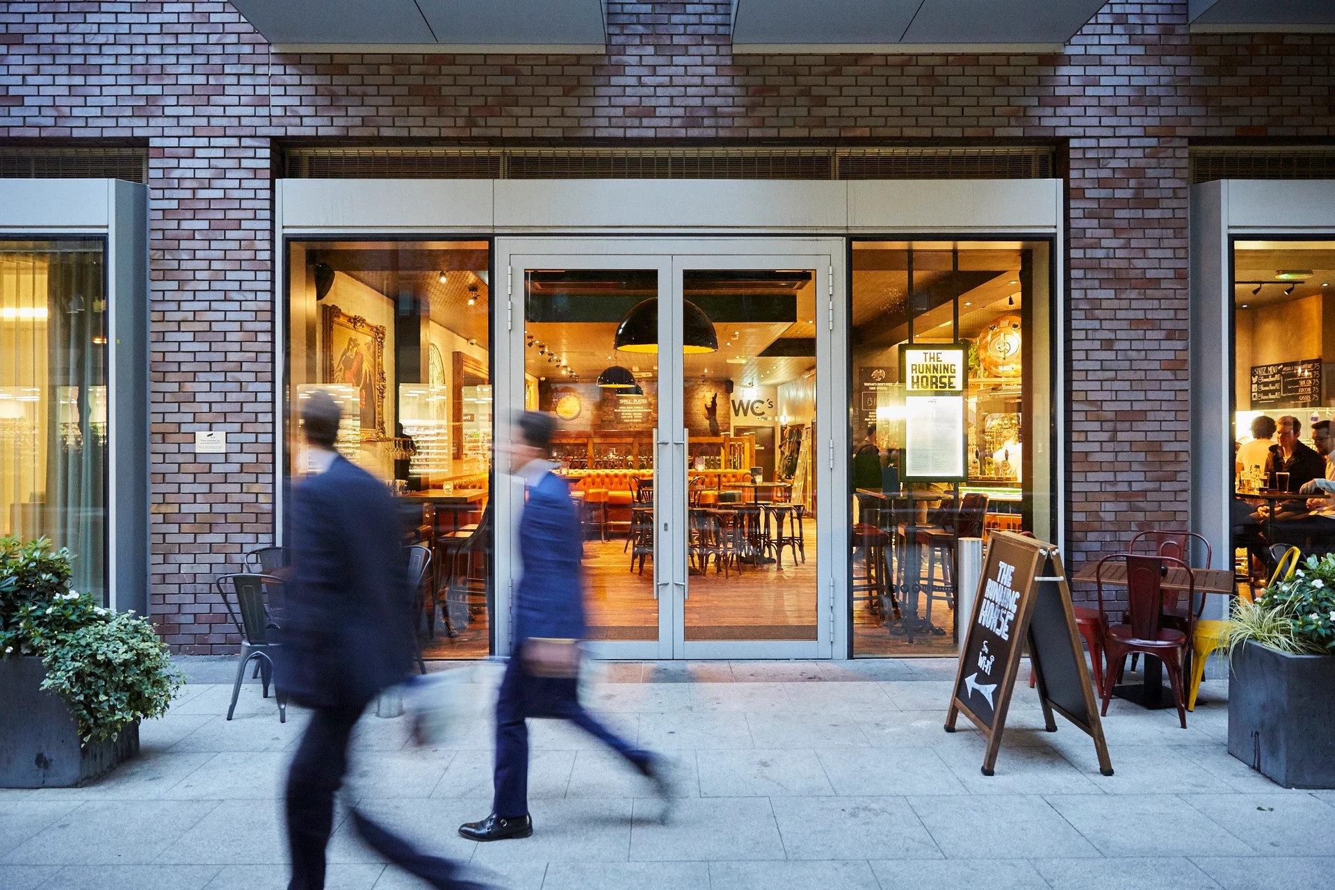 A brick building with large windows and a glass door is the focal point of a bustling street scene. Two individuals walk past the entrance, while a wooden signboard and potted plant add to the urban atmosphere. The scene is bathed in warm, inviting light, creating a lively and inviting atmosphere.