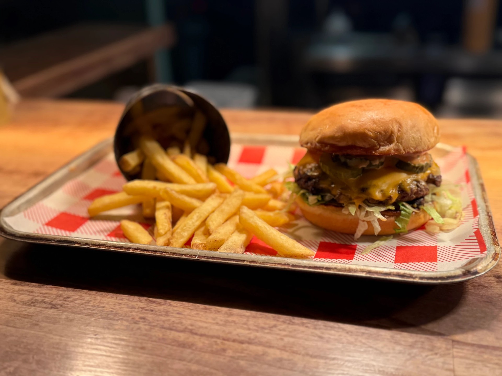 A golden-brown burger with a bun, lettuce, cheese, and tomato sauce is the centerpiece of a meal on a red and white checkered tray. A side of golden-brown fries is to the right. The tray is on a wooden table, and the background is blurred, emphasizing the meal. The image is a simple yet appetizing representation of a satisfying meal.