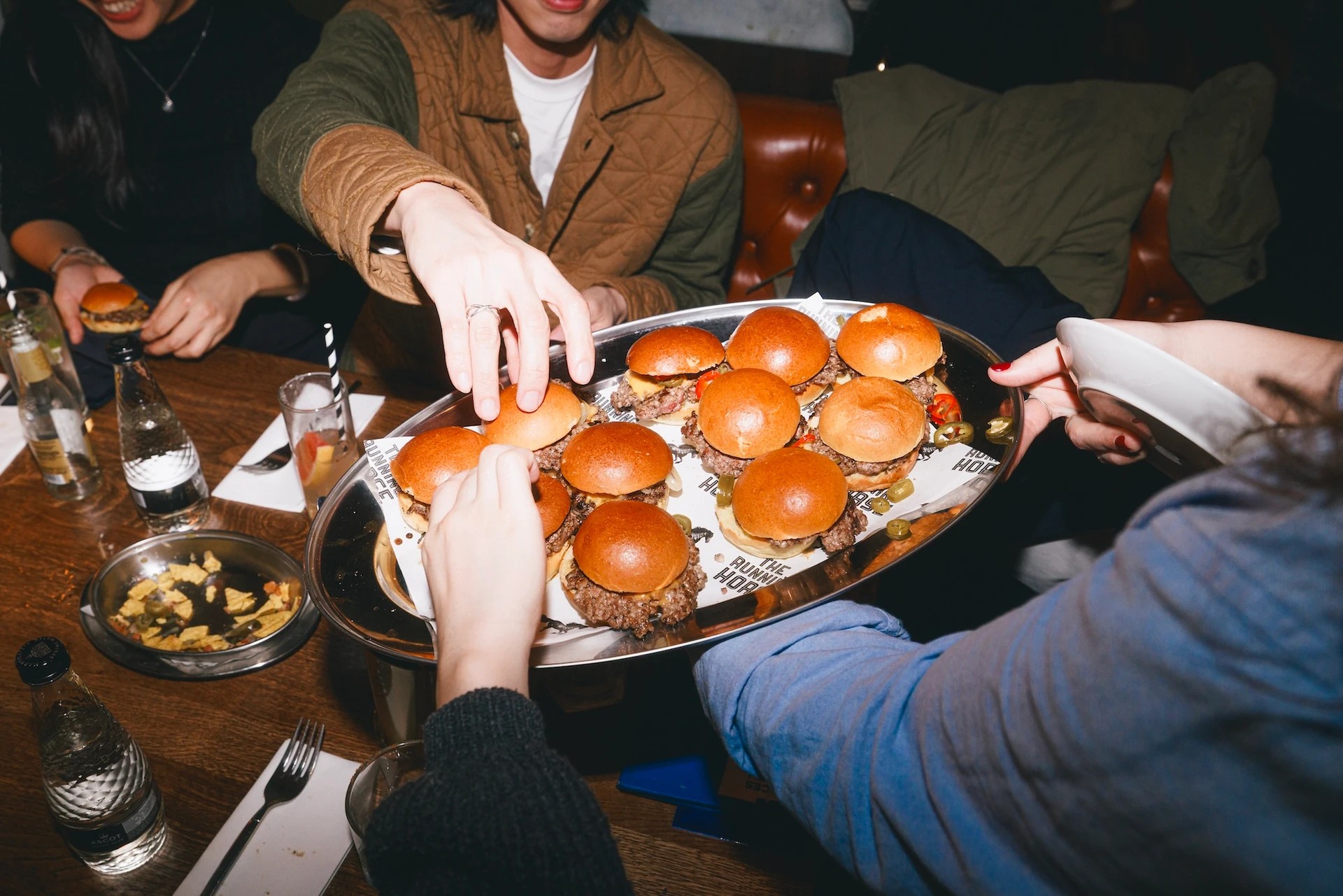 A lively gathering features a wooden table with a variety of food and drinks, including a tray of six golden brown burgers, a bowl of fries, a glass of water, a bottle of ketchup, and a glass of beer. A person in a blue sweater reaches for a burger, while another in a brown sweater holds a plate of food. The scene is filled with anticipation and camaraderie, with a warm and inviting atmosphere.