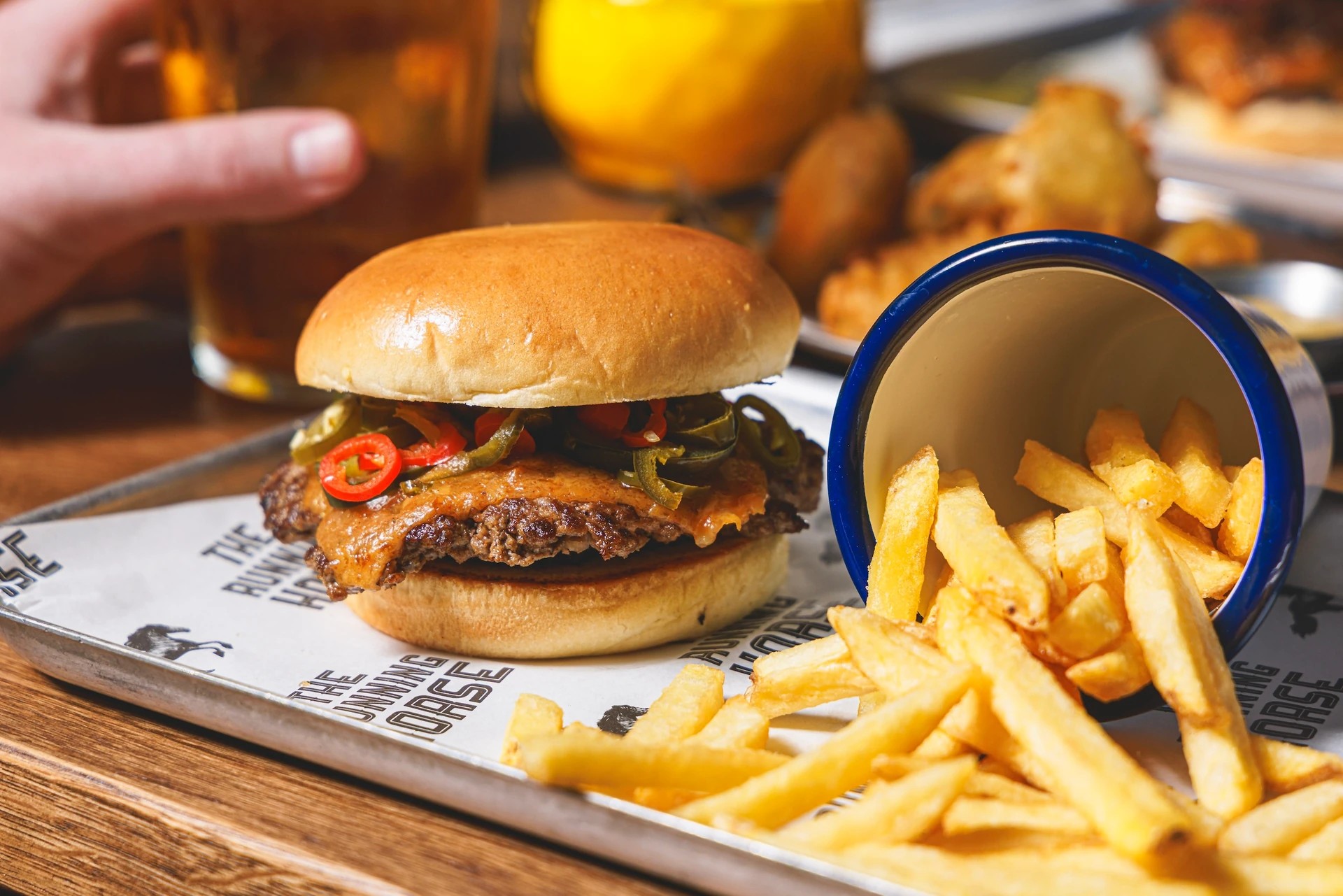A hearty meal is displayed on a wooden table, featuring a large burger with a brown bun, grilled chicken, and red peppers, accompanied by golden fries. A blue cup with a white lid holds a glass of beer, and a hand points at the burger. The scene is set against a black and white tray with black text, creating a visually appealing and appetizing meal.