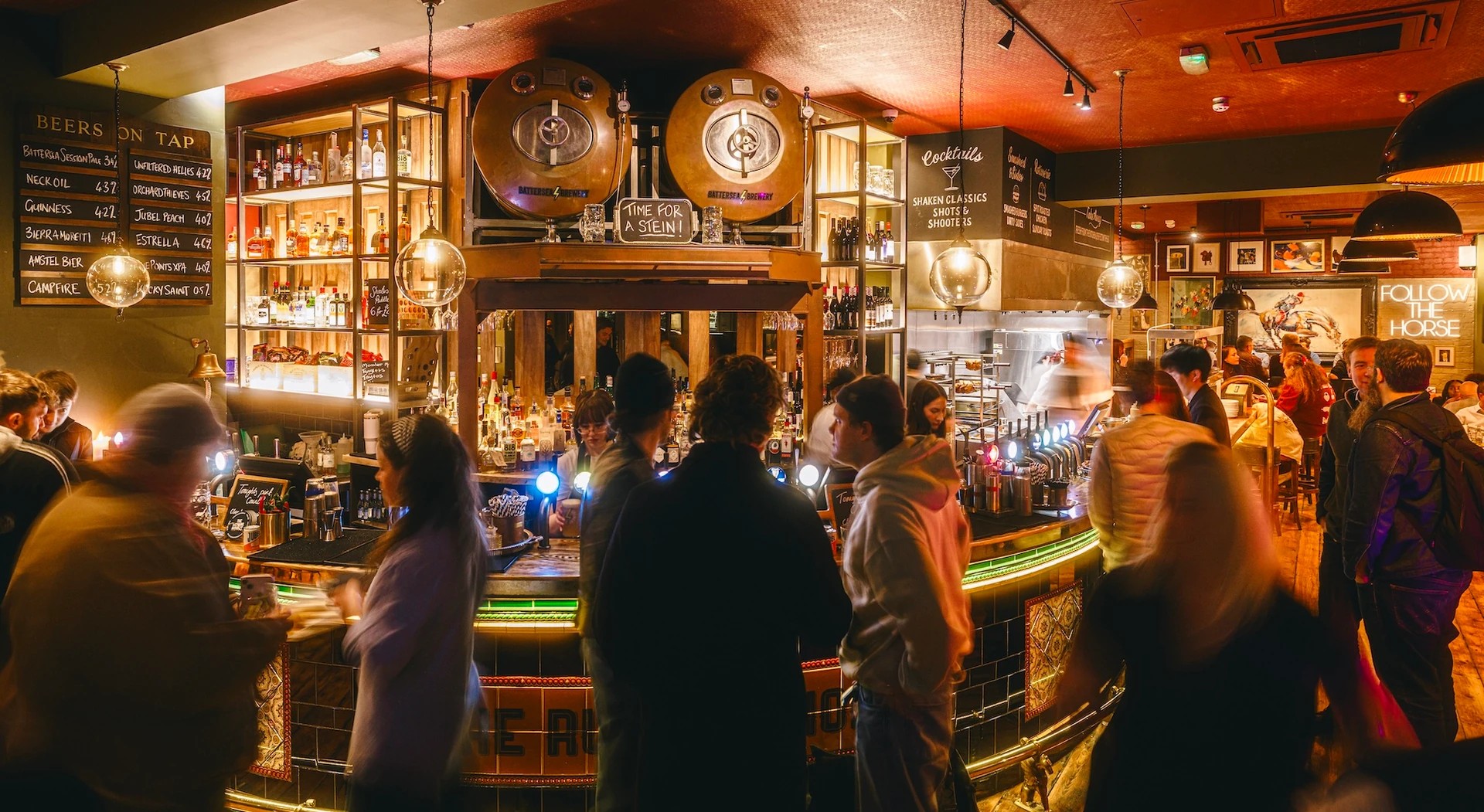In a dimly lit bar, a group of people gather around a wooden bar counter, with a large mirror reflecting the scene. The counter is adorned with various bottles and glasses, and the bar is filled with patrons. The warm, inviting atmosphere is enhanced by hanging lamps and a large clock. The perspective is from the front of the bar, looking towards the back, capturing the lively atmosphere of the establishment.