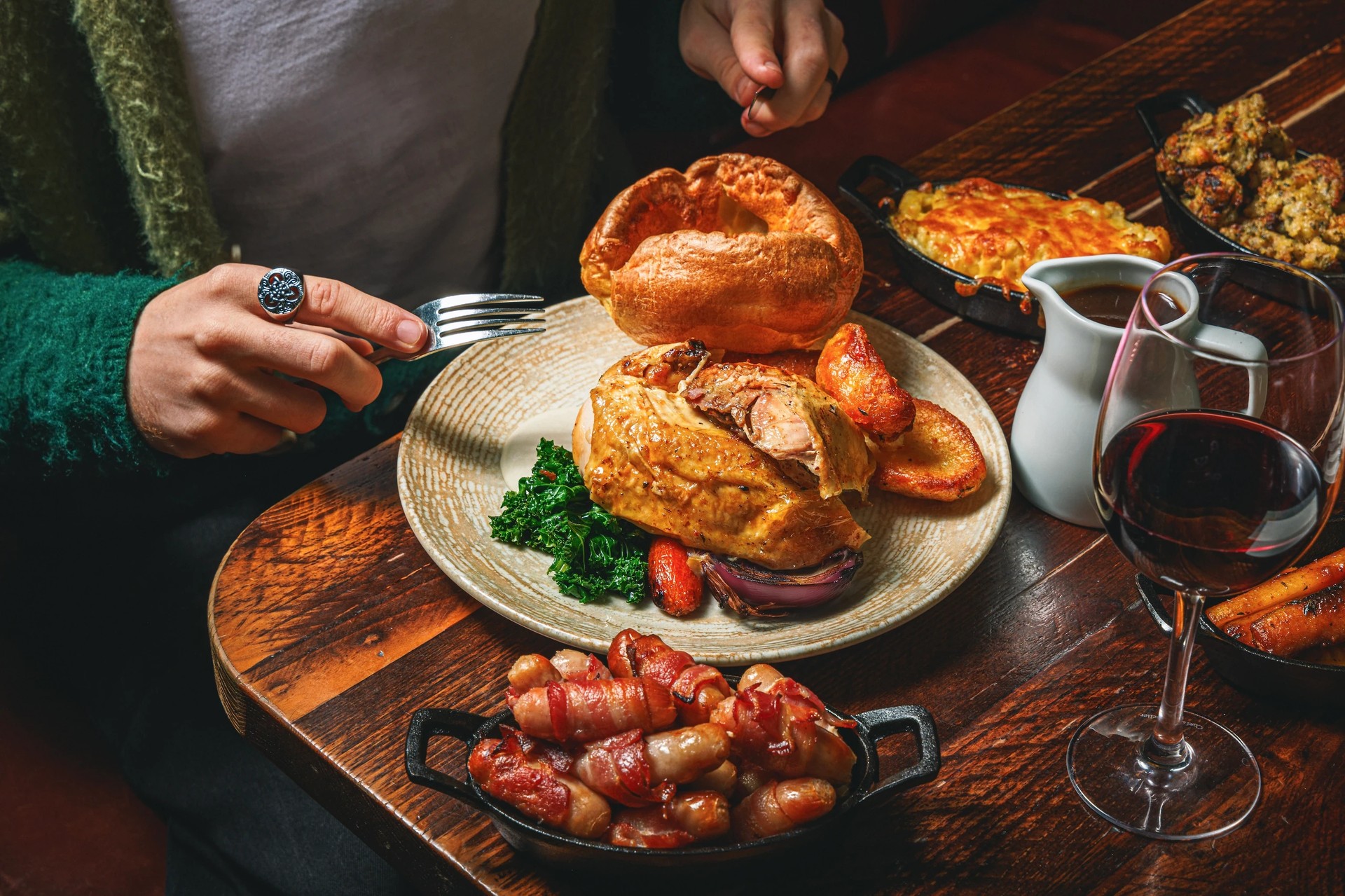 A person in a green sweater is seated at a wooden table, reaching for a large, golden-brown roast chicken sandwich with onions and red sauce. A plate of roasted potatoes and a pitcher of red wine are nearby. A white pitcher with a red liquid is also present. The scene is set against a dark background, emphasising the main subject and its surroundings.