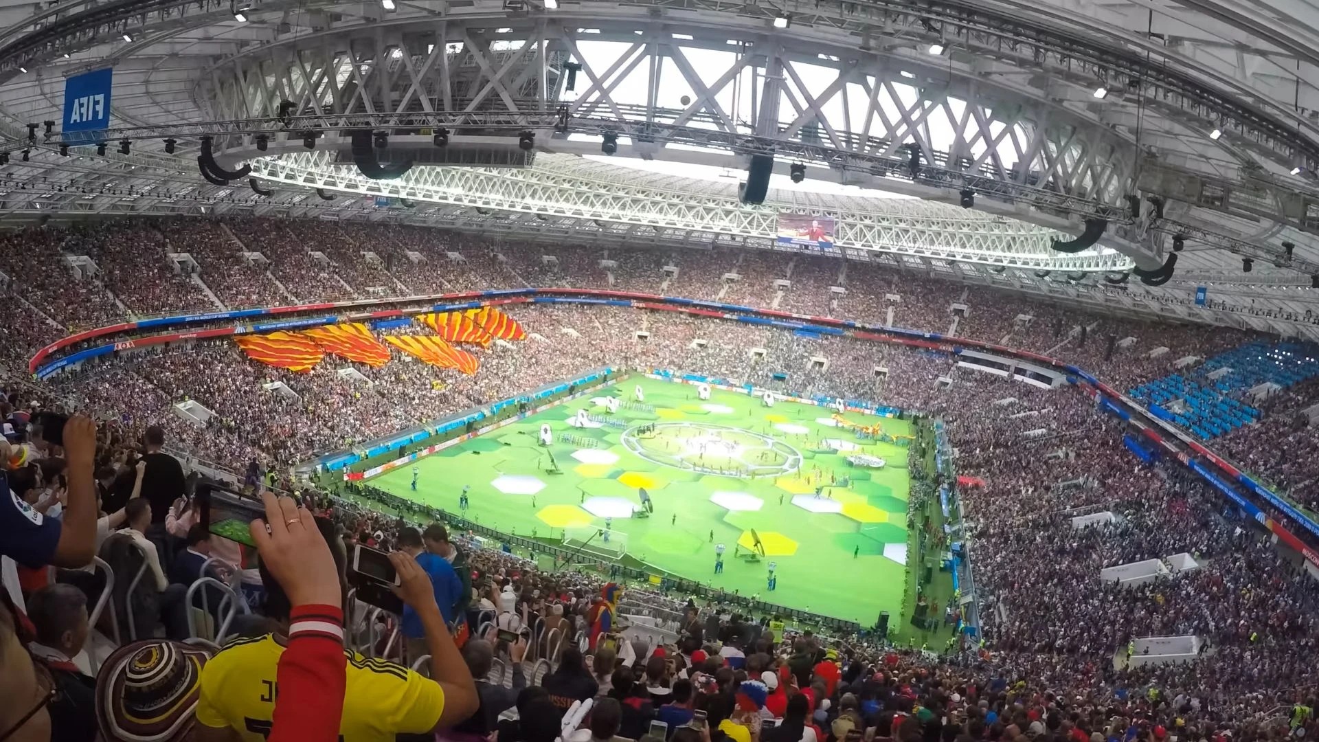 A large, well-maintained stadium filled with excited spectators is captured from a high vantage point, showcasing the vibrant green field with colorful flowers and a large banner. The stadium is surrounded by a white arch, and the image is taken during daylight, highlighting the energy and excitement of the event.