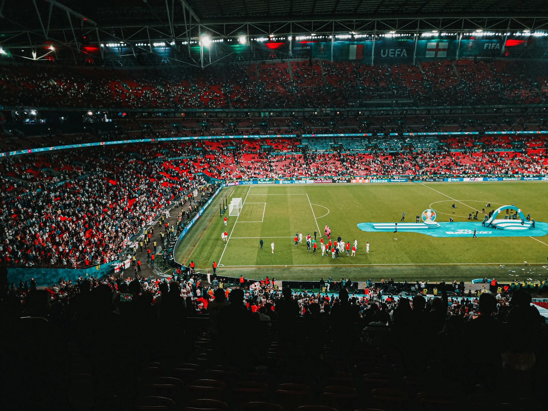 A nighttime soccer match is captured from a high vantage point, showcasing the vibrant green field, white goalposts, and red and white players. The crowd, filled with anticipation, is visible in the background, and the atmosphere is electric. The image is rich in detail and color, with no discernible text.