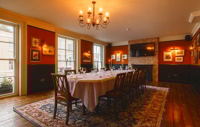 The image shows a luxurious dining room with a long table set for six, surrounded by wooden chairs and a rug. The table is adorned with a white tablecloth and wine glasses, and the room features a fireplace, framed pictures, and a chandelier. The walls are painted red, and the floor is made of wood. The room is well-lit, creating a warm and inviting atmosphere.