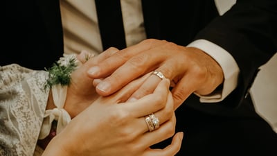 A close-up image captures a moment of intimacy between a bride and groom, with their hands intertwined in a romantic gesture. The bride wears a white lace dress and the groom dons a black tuxedo. Both hold wedding rings with diamond and gold accents, symbolizing their commitment to the union. The background is blurred, emphasizing the focus on the couple and their shared emotion.