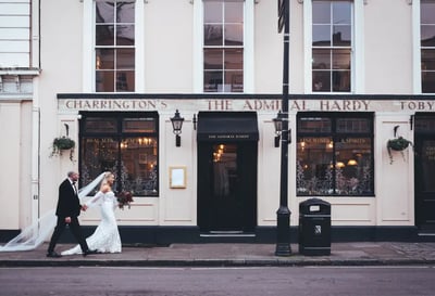 A newlywed couple in wedding attire walks hand in hand past a white "The Almance" pub in London, with a black trash can nearby. The scene is set against a backdrop of a street with a building and a street lamp.