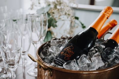 A black wine bottle with an orange label and a gold handle is in a copper ice bucket on a white tablecloth, surrounded by empty glasses. The background features a white vase with white flowers, creating a sophisticated and elegant atmosphere.