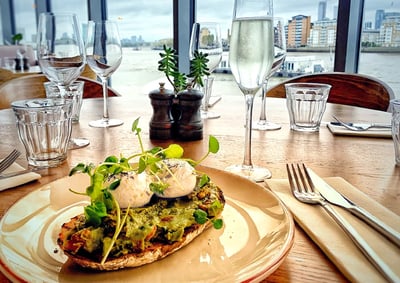 The image depicts a serene dining scene with a wooden table set for two, featuring a large, golden plate with a sandwich topped with green vegetables and a poached egg. Silverware is arranged on a white napkin, and a glass of water is nearby. A small plant in a black and white salt shaker adds a touch of nature. The city skyline is visible through the window, creating a harmonious balance between the indoor and outdoor elements.