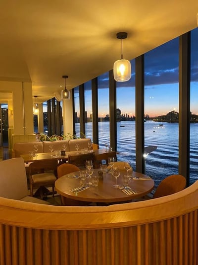 The image shows a luxurious restaurant with a curved dining area, featuring a wooden table with elegant chairs and a white tablecloth. The table is set with silverware and wine glasses, and is surrounded by a green sofa and a white coffee table. The restaurant is illuminated by hanging lights and a chandelier, with a serene view of a body of water and city skyline visible through the large windows. The layout is designed for comfort and elegance, with a harmonious color palette.