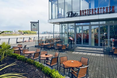 The image shows a modern, glass-facade restaurant named "Café de la Cité" with a wooden facade and metal roof. The restaurant is located on a street with a sidewalk and a sidewalk café. The café has tables and chairs arranged for patrons, and a street sign is visible in the background. The sky is overcast, casting a soft light on the scene.