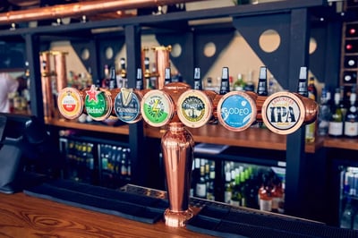 A copper beer tap with six distinct labels stands on a wooden counter in a bar, surrounded by shelves of beer bottles. The tap is centrally positioned, with the labels arranged in a row, inviting customers to choose their preferred beer. The background features a black ceiling and shelves of beer bottles, creating a well-organized and inviting atmosphere.