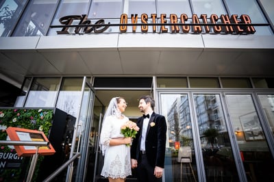 A newlywed couple, dressed in elegant attire, stands outside a modern hotel entrance, with the bride holding a bouquet of flowers and the groom in a black suit. The hotel has a glass facade, a large window, and a sign indicating a 100% refund policy. The scene is set against a backdrop of lush greenery and a building with a large window.