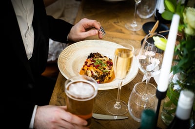 A man in a black suit is seated at a wooden table, ready to enjoy a meal. The table is set with a white plate of food, a gold wine glass, a silver fork, and a white vase with a bouquet of flowers. The scene is set for a formal dining experience.