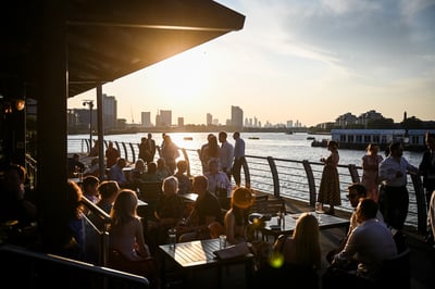 The image captures a lively outdoor scene at sunset, featuring a bustling restaurant with patrons enjoying the view of the city skyline and a river. The restaurant has wooden tables and chairs, and the atmosphere is filled with conversation and relaxation. The city skyline is visible in the background, with a boat on the river adding to the scene's charm. The image is rich in detail and color, with a warm, inviting ambiance.